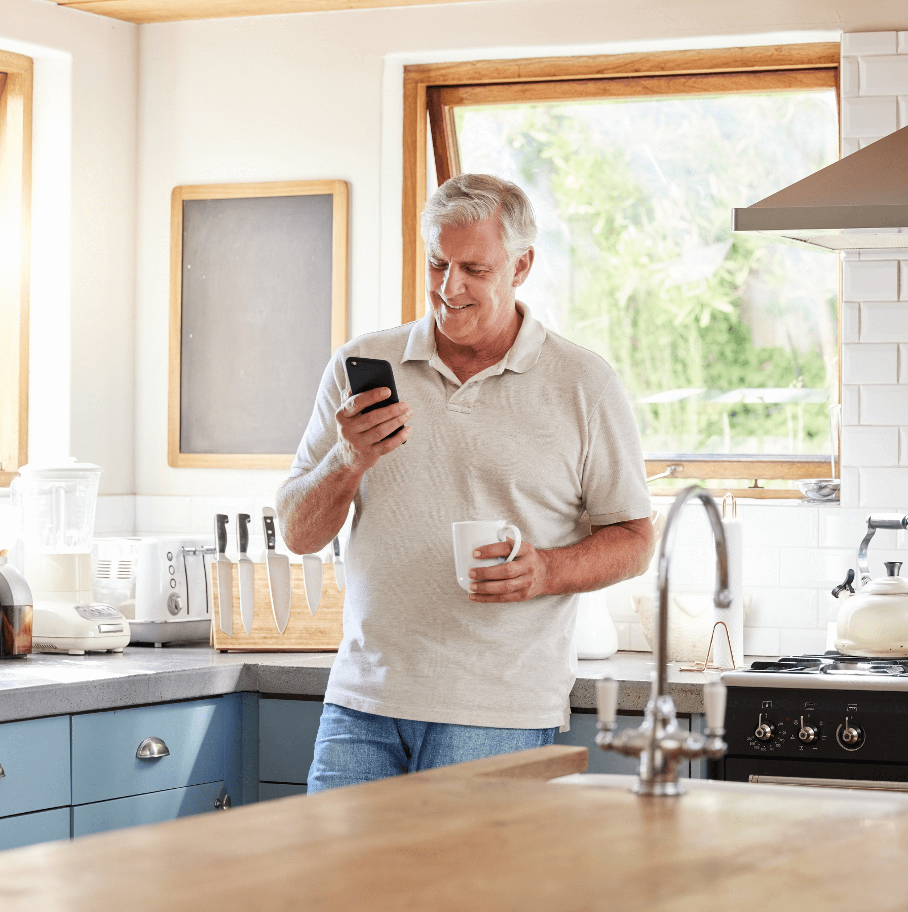 A man in a kitchen holding a mug and a smartphone.