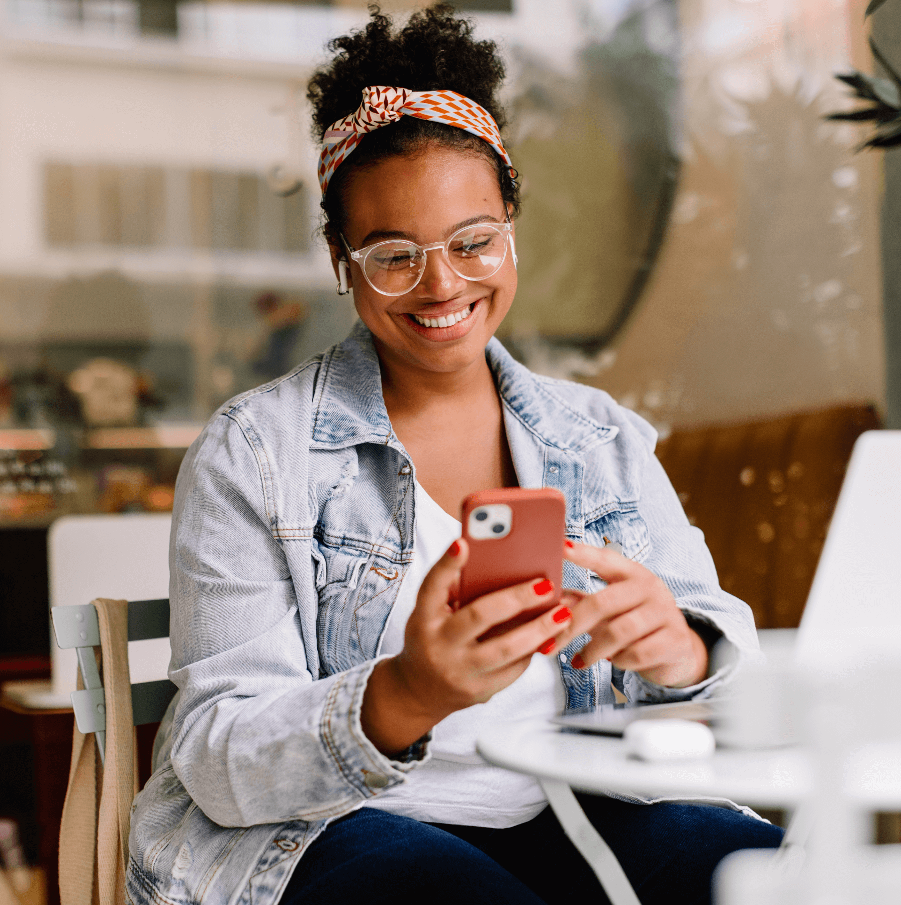Person in a denim jacket smiling at a smartphone while sitting at a cafe table.